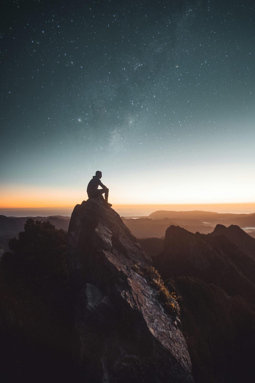 A lone figure sitting on a mountain summit at dawn beneath a star-filled sky