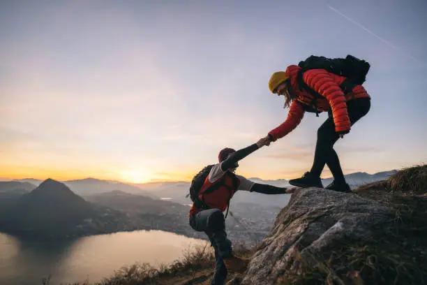 One hiker reaching down to help another up a mountain ridge at sunset